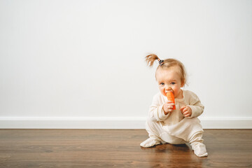 a baby in a milk-colored jumpsuit eats a carrot at home against a white wall, the first teeth are being cut, the baby is scratching the gums