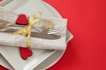 Festively served table for Valentine's Day with a plate, fork and knife, red hearts and ribbon on a red background