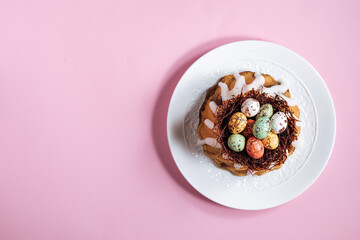 Easter cake with colorful eggs in a chocolate nest on pink background and flowers. Happy Easter concept. Dessert also known as  Kuglof. Top view