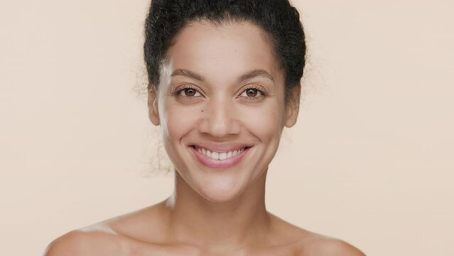 Close-up beauty portrait of gorgeous slim African American young woman with dark curly hair lifts eyes with fingers smiling wide for the camera on beige background | Droopy eyes removal concept