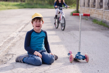Young boy crying, sitting on the road after falling from his push scooter