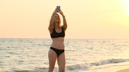 beautiful slender young blonde woman in black swimsuit holds phone in her hands and make selfie. Photo of yourself against backdrop of sunset on sea or ocean on beach.