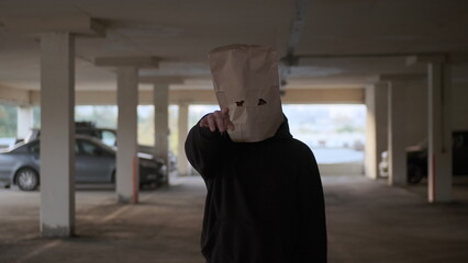 character with paper bag on his head in black hoodie points finger at you. You are in deep trouble. scary masked boogeyman threatens you while standing in car parking lot.