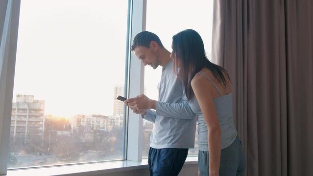 Young man is texting with his mistress at home by window. Wife approaches her husband and takes phone to see correspondence. Untrustworthy family relationships.