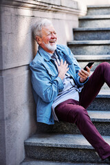 A happy senior student is sitting on the stairs with a phone in his hands and takes a break between...
