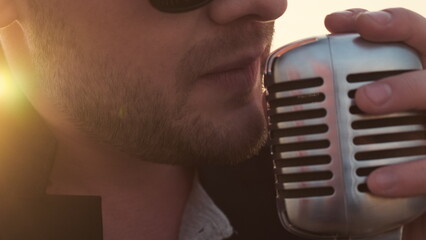 bearded rock star singer with glasses sing expressive song in retro microphone. Extreme close up face man with stubble or beard in black jacket, holding microphone with hand and yelling at it.