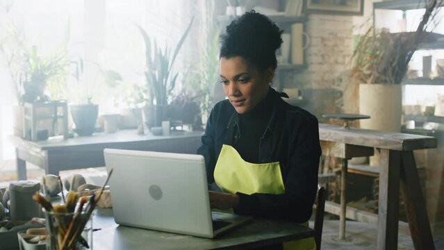 Young pretty woman potter is sitting at table in pottery studio, opens laptop to work on design of new ceramic items, looking at camera, Foreground, Slow motion.