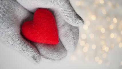 Close-up of hands in gray knitted warm mittens holding a red soft heart against the background of lights,blurs,copy space.Valentine's day concept.Beautiful holiday card.Love.Winter.Romance.