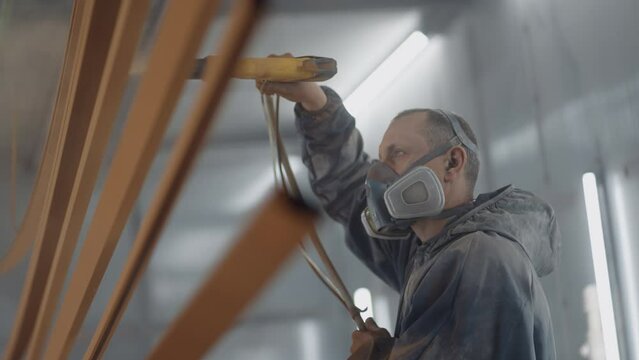 A man in a respirator is painting metal with a powder method. Painting shop. The painter works with a spray gun paint is applied