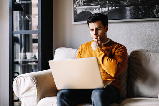 Smiling hispanic millennial man relax on comfortable couch in living room work on laptop gadget, happy biracial young male freelancer relax on sofa at home using computer, technology concept