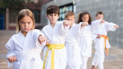 Fototapeta na wymiar Concentrated tween girl in white kimono exercising new techniques during group outdoors taekwondo lesson in schoolyard on warm day
