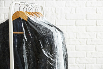 Rack with clean jackets and shirts in plastic bags on white brick background, closeup