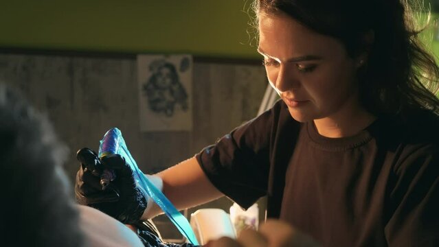 Close-up of young adult female tattooer, tattoo artist works with customer shaved chest. Woman tattooist master in hand gloves fills ink dye with protected needle machine in male clients body chest.