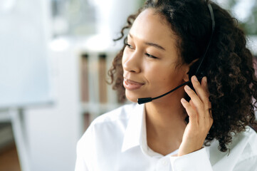 Close-up of black curly haired young woman with headset, female african american support service worker, call center operator, in modern office, looking away, smiling