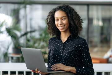 Fototapeta na wymiar Portrait of curly attractive positive african american pleasant young business woman, freelancer, real estate agent or manager, holding open laptop in office, looking at the camera, smiling friendly