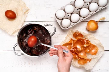 Keuken foto achterwand Easter eggs, the process of coloring with natural dye, onion husks in a small saucepan on a white wooden background and a woman's hand holding a colored egg in a spoon © Надежда Урюпина