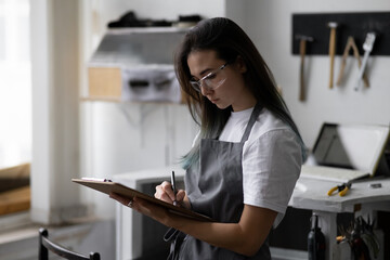 Young woman jeweler in gray apron holds clipboard in her hands. Makes sketches for new products....