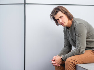Millennial man with long hair and tense look sits near grey wall. Serious man in earth toned casual...