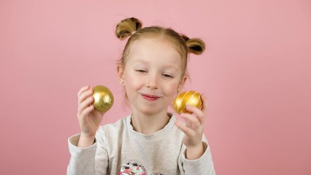Funny little blonde girl smiling and playing with golden easter eggs on pink background. Happy Easter concept