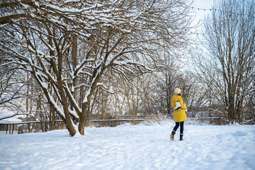 Young woman in yellow jacket with ice skates walking through snowy park. View from behind.