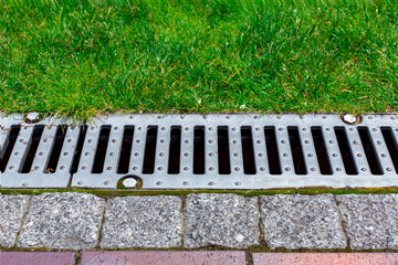 gray grating of the drainage system for drainage of rainwater in the park at edge of sidewalk from granite tile with green lawn, landscaping a public garden close up view, nobody.