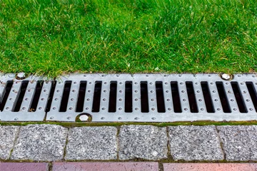 Fotobehang gray grating of the drainage system for drainage of rainwater in the park at edge of sidewalk from granite tile with green lawn, landscaping a public garden close up view, nobody. © Александр Беспалый