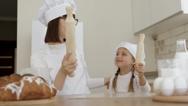 Mother or older sister and little kid daughter or younger sister in hats of chef have fun laughing and fighting on rolling pins and teaches how to bake pastries or homemade bread together