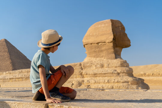 The boy in hat sits in front of the sphinx and looks at it. 
