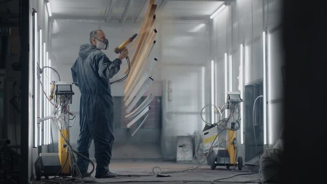 A man in a protective suit and mask sprays paint through a spray gun in slow motion on steel parts.