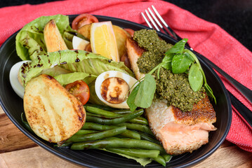 A serving of salmon steak, with basil pesto, green beans, vegetables and salad in an oval shaped bowl. Side view.