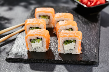 Maki sushi on dark stone table. Philadelphia maki with salmon. Sushi roll with cheese, cucumber inside, salmon outside. Style concept japanese menu with black background, leaves and hard shadow.