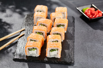 Maki sushi on dark stone table. Philadelphia maki with salmon. Sushi roll with cheese, cucumber inside, salmon outside. Style concept japanese menu with black background, leaves and hard shadow.