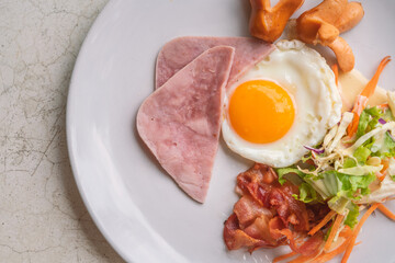 Top view easy breakfast menu, fried egg, ham, bacon, sausage, and salad. Breakfast on white plate on white stone table, natural light.