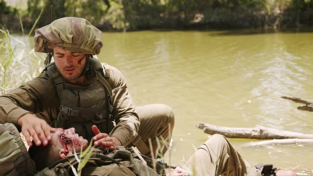 Soldier washes head of badly injured teammate with a large red wound on his face at a dirty water pool. Close up shot