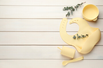Flat lay composition with silicone baby bib and plastic dishware on white wooden background. Space...