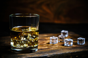 Glass chilled whiskey with ice cubes on wooden background in cellar.
