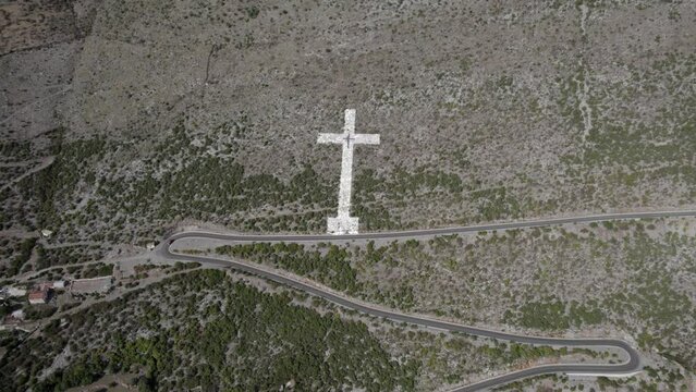 Drone video of descriptive panning over the giant white cross at the beginning of the mountain pass of the Sh20 road in Bridje, Albania, houses of the village can be seen, winding road.