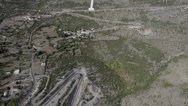 Drone video of descriptive panning over the beginning of the mountain pass of the Sh20 road in Bridje, Albania, we see the religious white cross, houses of the village can be seen, winding road