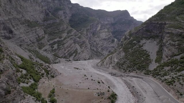 Drone video of a diagonal plane discovering the Sh20 highway between the mountains and the Cemi river in Albania at the height of Selce, cloudy sky and winding road.