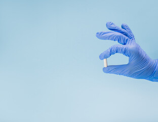 The hand of a caring doctor in a blue latex glove holds one white pill or tablet on a light blue background with a place for text, disease prevention