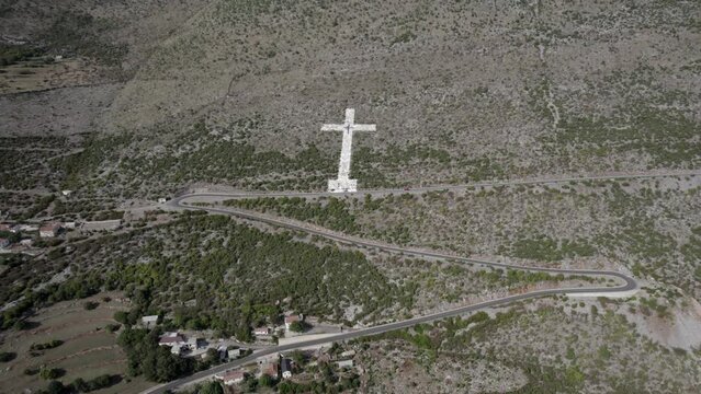 Drone video of frontal plane towards the giant white cross at the beginning of the mountain pass of the Sh20 road in Bridje, Albania, houses of the village can be seen, winding road.