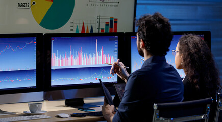 Closeup shot of big center monitor showing company analysis target circle graph and chart report in trading room full of computer screen while unrecognizable unknown male and female staff working