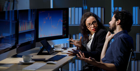 Fototapeta na wymiar Caucasian male stock investor discussing with African American female businesswoman sitting in trading room with graph chart analysis screens and monitors.