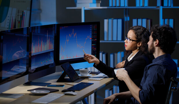 Caucasian male stock investor  discussing with African American female businesswoman sitting  in trading room with graph chart analysis screens and monitors.