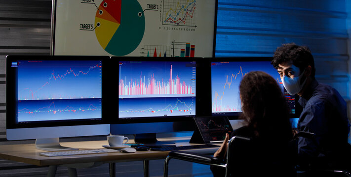 Caucasian male stock investor  discussing with African American female businesswoman sitting in wheelchair wearing face mask in trading room with graph chart analysis screens and monitors.