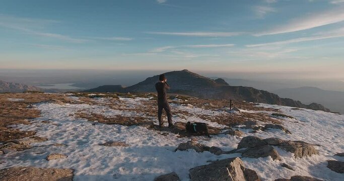 Back view wide angle shot of young hiker taking photos with his camera on summit of snowy mountain in Guadarrama mountains, Madrid, Spain.