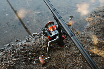 Top view spinning rod, red steel reel on shore near water outdoors. Fishing hobby, sport and leisure concept