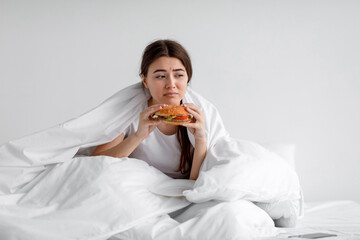 Sad upset depressed european millennial woman eating burger in bed under duvet at home alone