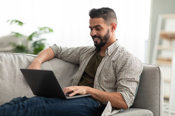 Freelancing, online job or studies. Cheerful young Arab man sitting on couch with laptop, working or learning from home