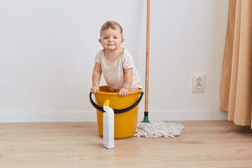 Indoor shot of sad upset little cute baby girl of one year age standing in yellow bucket near mop...